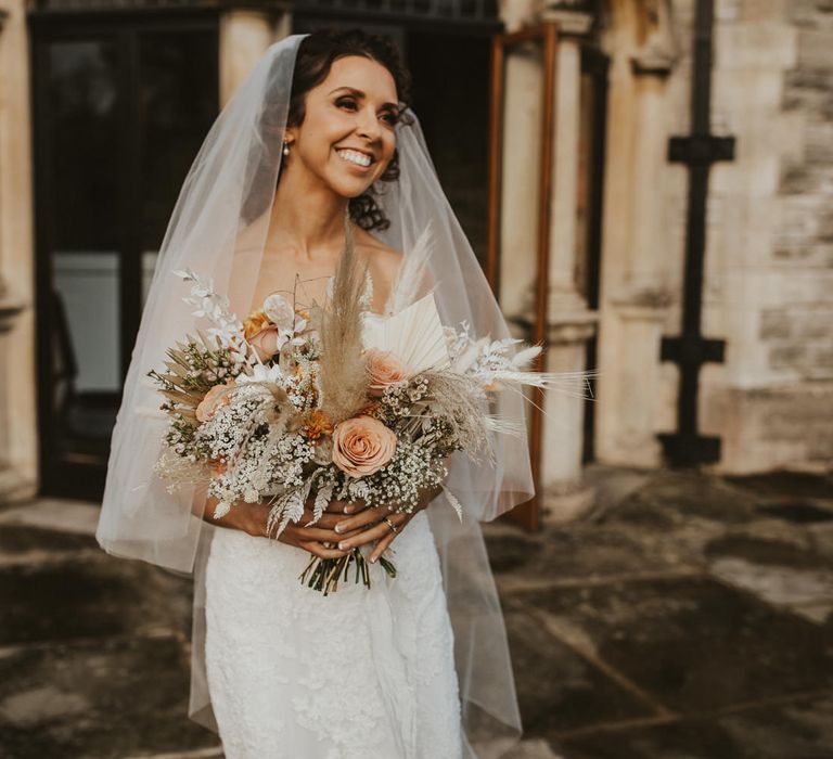 Bride in lace Pronovias wedding dress and veil holding a peach and neutral fresh and dried flower bouquet 