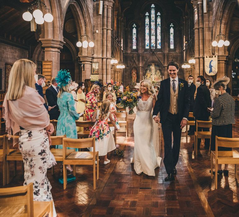 Smiling bride in Justin Alexander wedding dress walks back down the aisle in church ceremony holding hands with smiling groom