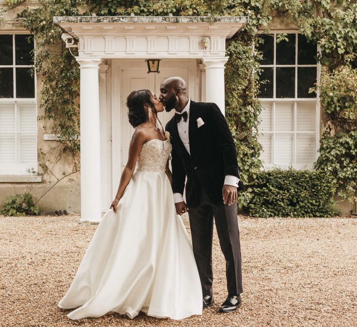 Black bride in a strapless Oleg Cassini wedding dress with beaded bodice kissing her groom in a velvet tuxedo and bow tie outside Northbrook Park 