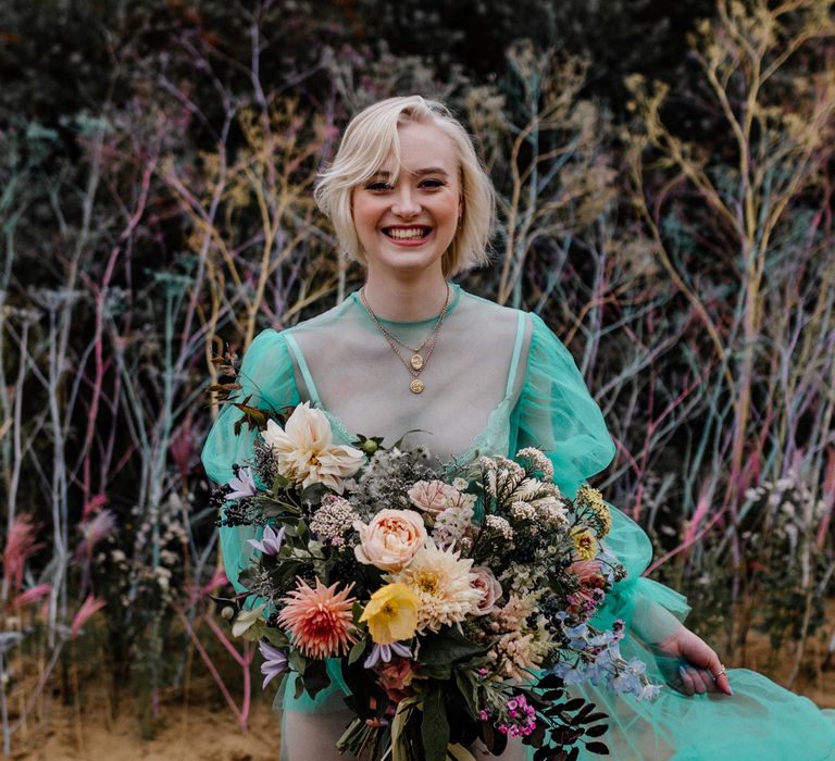 White blonde bride with bobbed hair in a sheer green pastel wedding dress holding a colourful wedding bouquet tied with ribbon