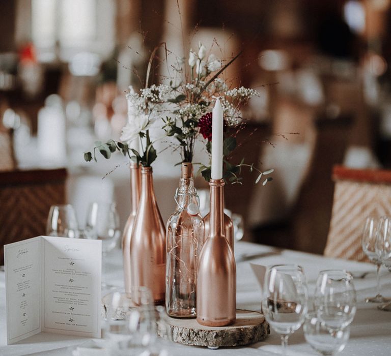 Rose gold bottles with white candles, gypsophila and copper string fairy lights on rustic bark board for wedding table decor