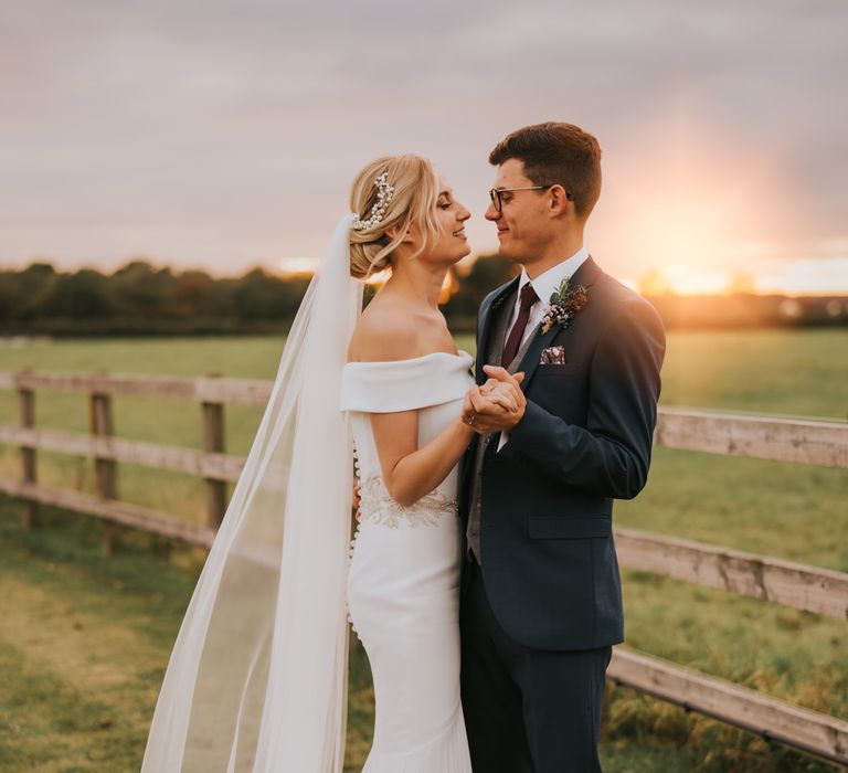 Bride & groom look lovingly at one another in the countryside as the sun sets in the background