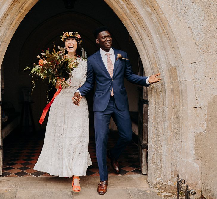 Bride in a Broderie Anglaise wedding dress and flower crown and groom in a navy suit exiting the church, just married 