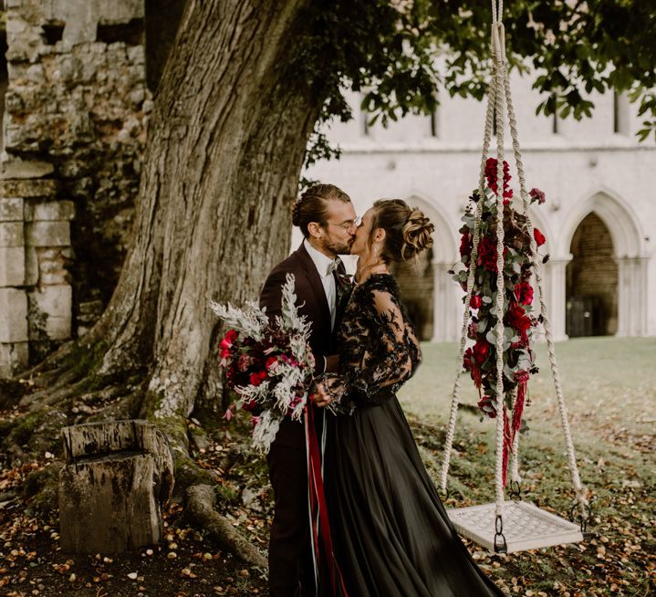 Bride in a black wedding dress with lace bodice kissing her groom in a burgundy suit by a tree and tree swing 