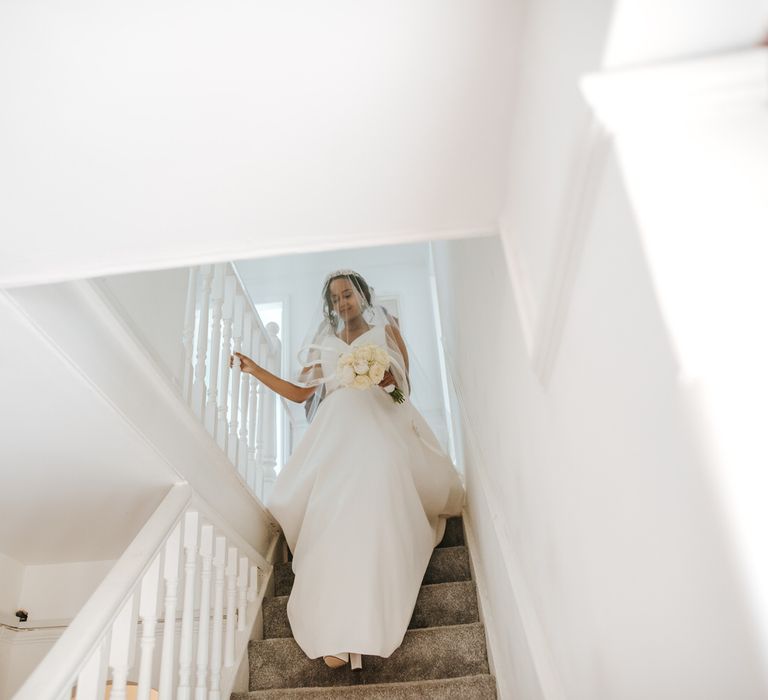 Bride in a sateen wedding dress holding a white bouquet walking down the stairs on the wedding morning 