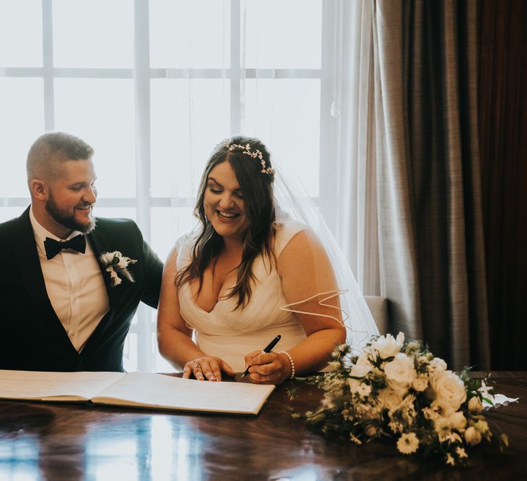 Bride & groom sit together during their wedding ceremony and sign marriage certificate in front of window