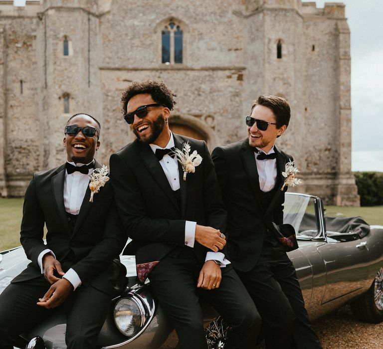 Happy groom and groomsmen in three-piece black suits with dried flower buttonholes laughing whilst sitting on the bonnet of the classic wedding car 