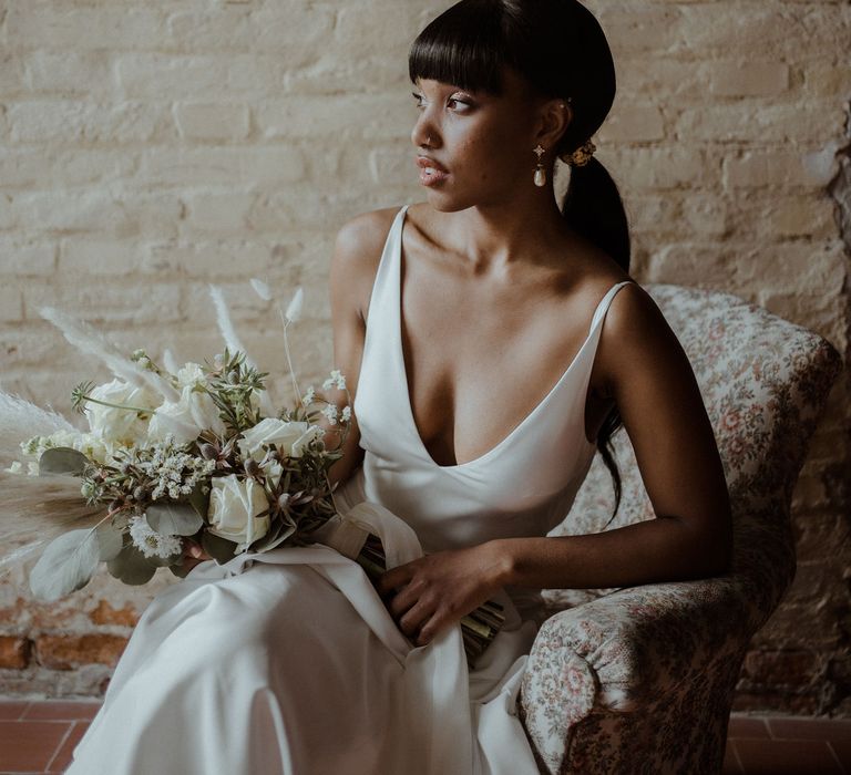 A Black bride sits on a cream chair holding a bouquet of dried flowers and grasses. She looks off to the side. She has pearl drop earrings and her hair tied back in a low ponytail.