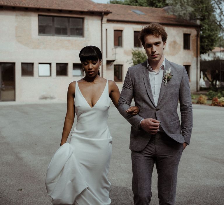 A bride and groom walk outside their venue. He wears a grey suit and she holds up the skirt of her long white dress.