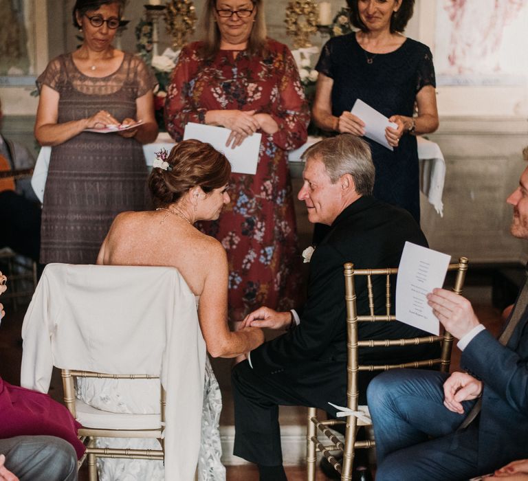 Bride & groom look lovingly at one another whilst sat down at the front of the Chapel as wedding party looks on