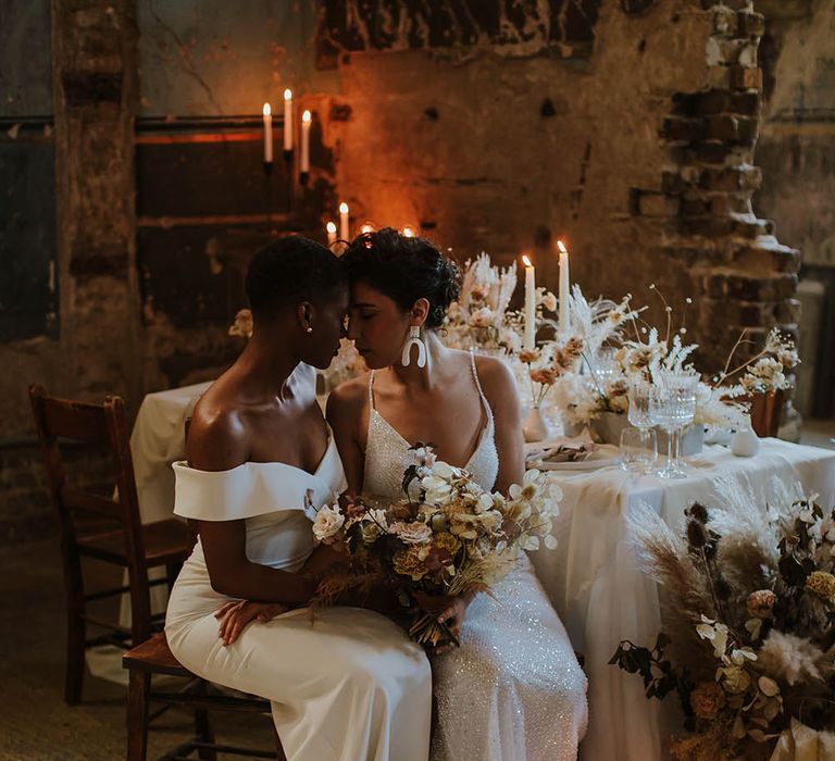 LGBTQI+ couple sitting at their wedding reception table in an off the shoulder wedding dress and sparkly gown holding a neural flower wedding bouquet 