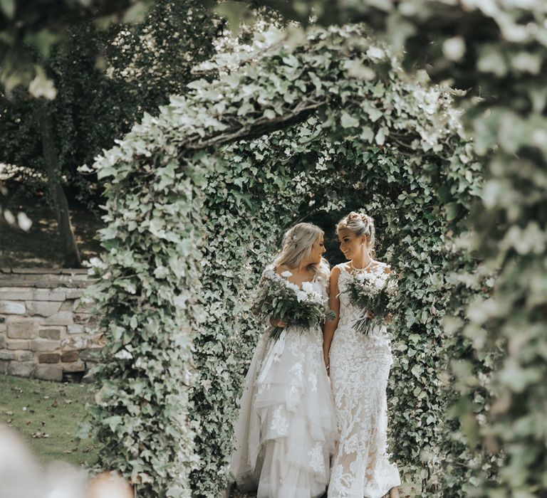 Two brides embrace under ivy covered arches at Crab & Lobster fairytale wedding.