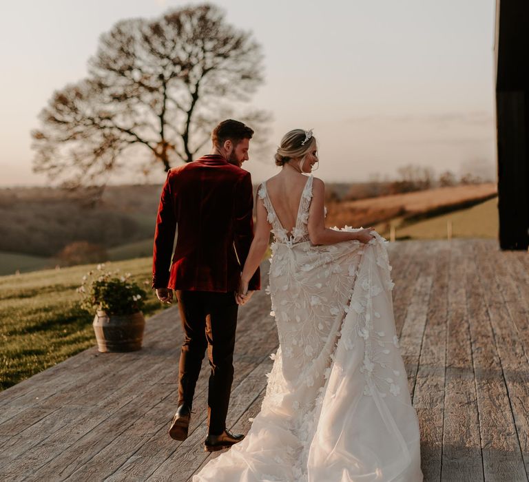 Bride in a princess appliqué wedding dress holding her grooms hand in a red velvet jacket on the decking at Botley Hill Barn