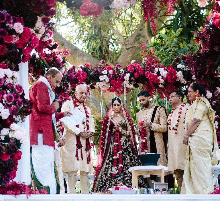 Bride & groom gather with family during outdoors ceremony surrounded by deep red florals