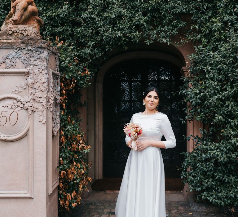 Bride stands within doorway surrounded by green foliage on her wedding day