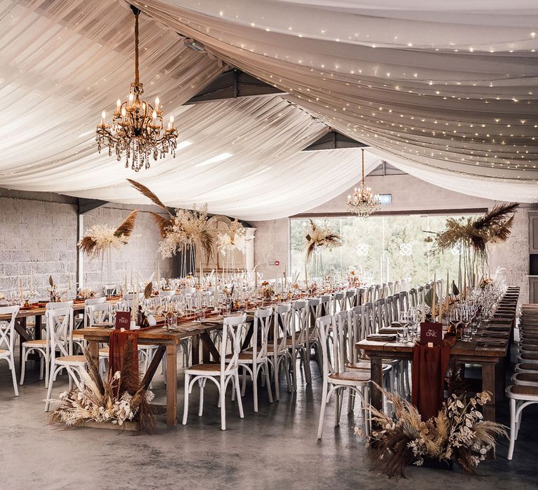 Boho wedding breakfast with long wooden table, burgundy table runner, pampas grass, white chairs and white drapery for summer wedding in Dorset