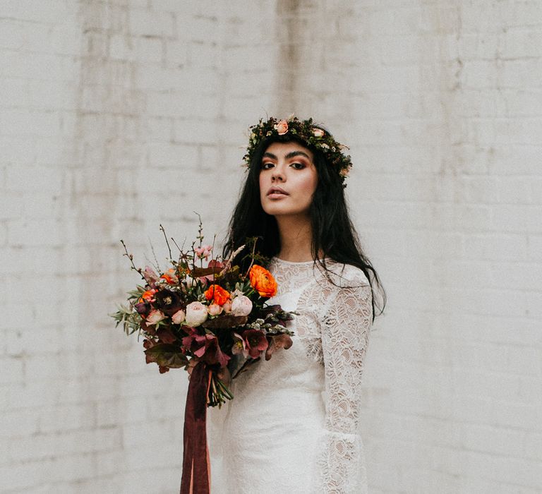 Boho bride in a lace wedding dress with long sleeves wearing a flower crown and holding a deep orange wedding bouquet tied with ribbon 