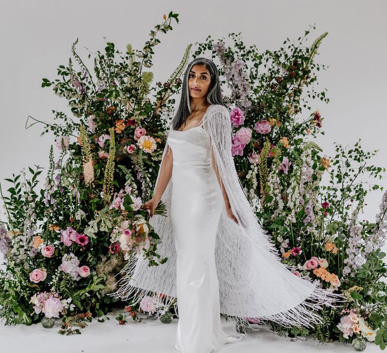 Bride in satin Halfpenny London wedding dress and tasselled bridal cape stands in front of multicoloured floral installation for wedding at Loft Studios London