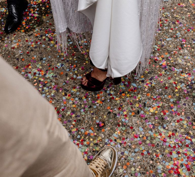 Groom in linen suit trousers and gold Nike Air Force 1 trainers stands next to bride in satin Halfpenny London wedding dress and tasselled bridal cape on floor on multicoloured confetti during wedding reception at Loft Studios London