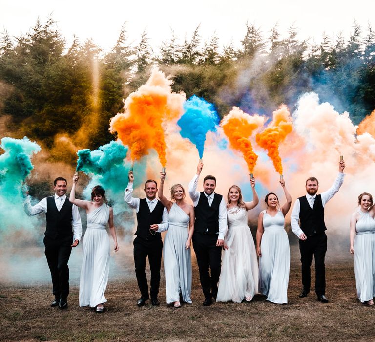 Wedding party photo with bridesmaids in grey dresses and groomsmen in black suits holding orange and green smoke bombs 