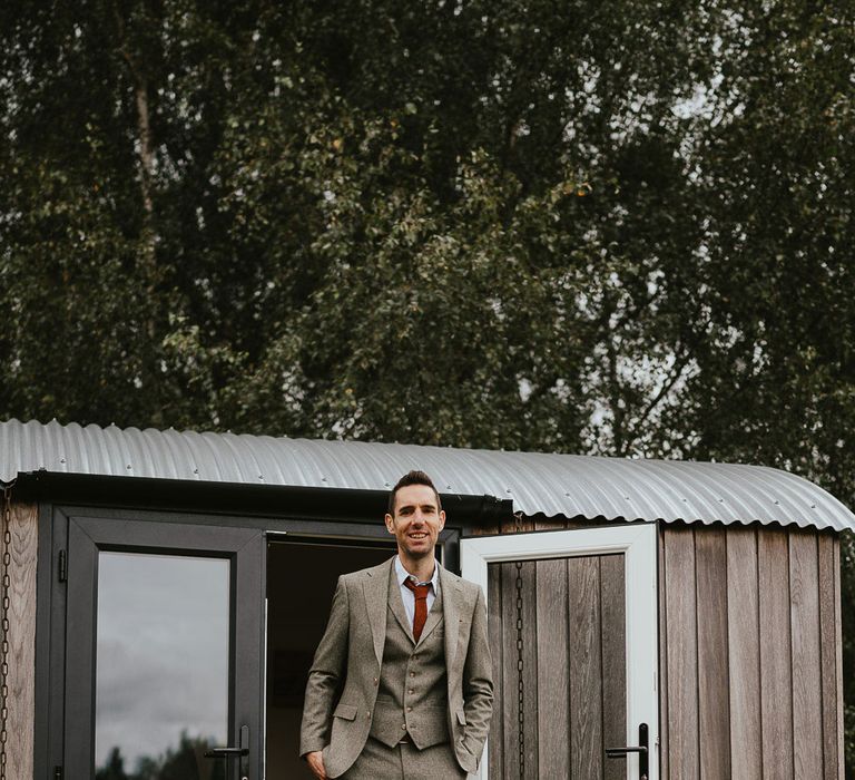 Groom in light three piece suit, red tie and brown shoes stands on doorstep of wooden Shepards hut before late summer wedding in Norfolk