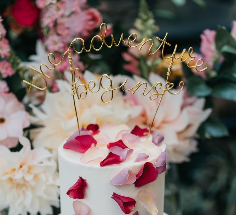 two tier buttercream wedding cake with edible petal decor and gold wire where the adventure begins cake topper 