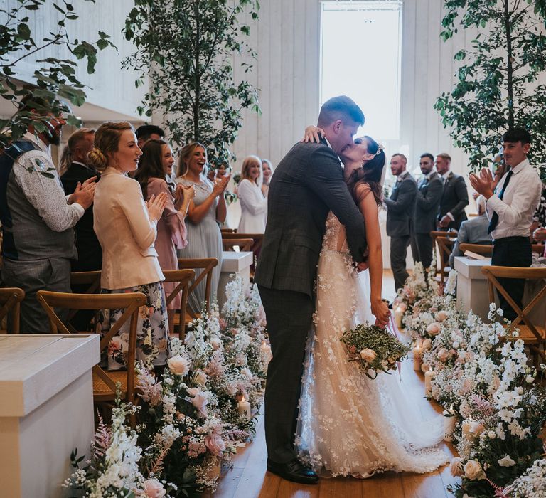 Bride in applique Berta wedding dress holding white and blush pink wedding bouquet kisses groom in grey checked quit as wedding guests clap during wedding ceremony at Primrose Hill Farm