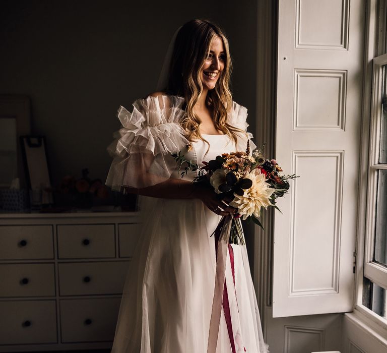 Bride in white Halfpenny London Mayfair dress with ruffle sleeves holding floral bouquet stands by window before church wedding ceremony in Cornwall