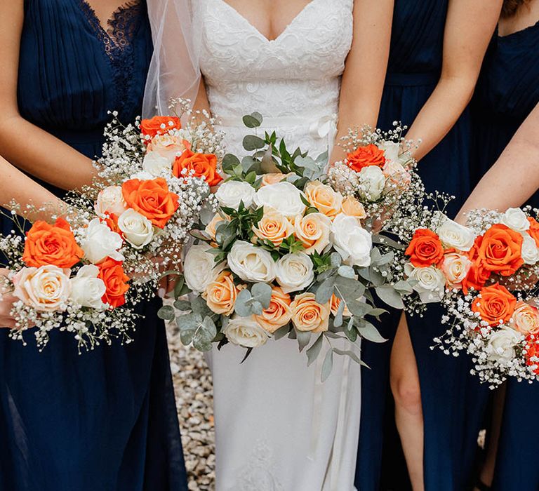 Bride stands with her bridesmaids who hold colourful orange and peach bouquets complete with green foliage