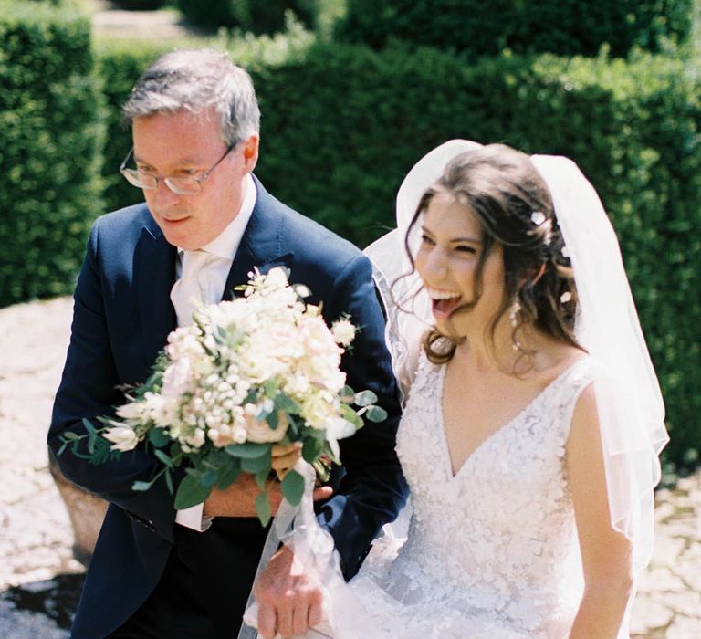 Bride walks down the aisle with her father whilst holding white floral bouquet