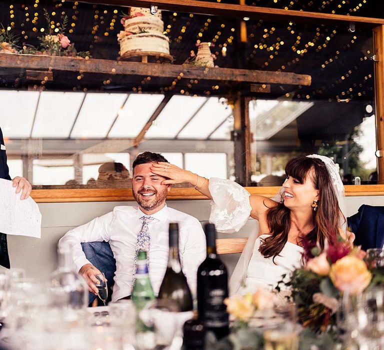 Bride in a strapless wedding dress with lace detachable sleeves feeling her grooms head as the best man delivers his wedding speech 