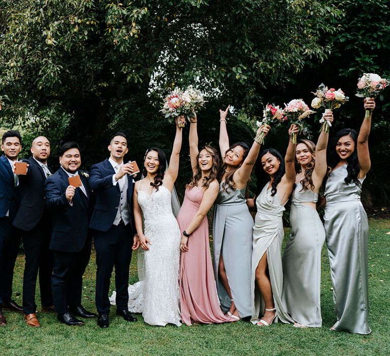 Wedding party picture with groomsmen in navy suits and bridesmaids in satin dresses holding their bouquets in the air