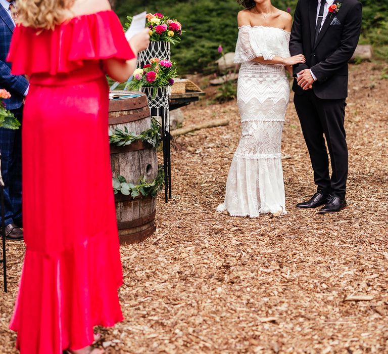 Bride in strapless Grace Loves Lace wedding dress and flower Cornwall stands with groom in black suit and bridesmaid in pink bridesmaid dress performs wedding reading during outdoor ceremony