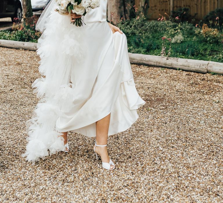 Bride in halterneck Halfpenny London wedding dress and veil with Jimmy Choo wedding shoes holds white and green bridal bouquet as she walks across driveway