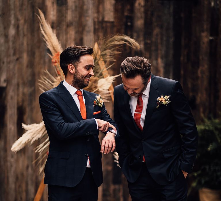 Groom in a navy suit with brown bridges and an orange tie standing at the wooden triangle altar decorated with pampas grass with his best man 