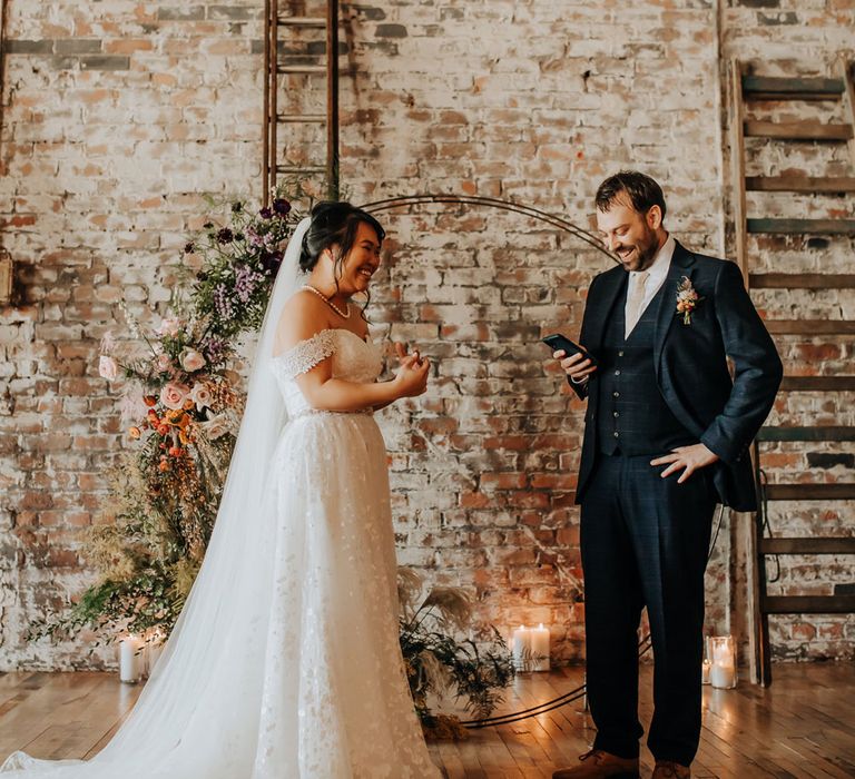 Bride in bardot wedding dress and veil stands by floral moongate whilst groom in blue suit reads vows off his phone at wedding ceremony in Liverpool