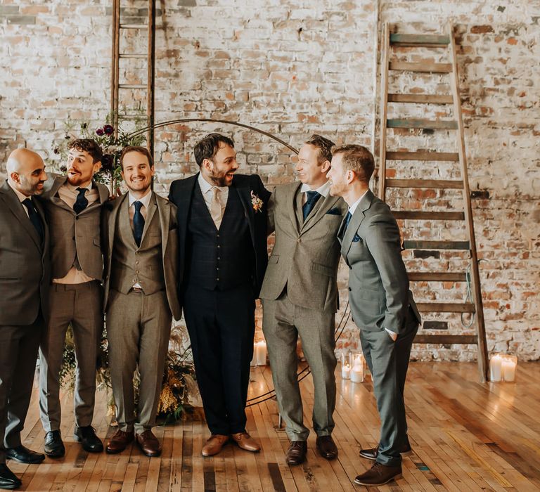 Groom in blue check three piece suit stands with groomsmen in grey three piece suits and blue ties in front of floral moongate