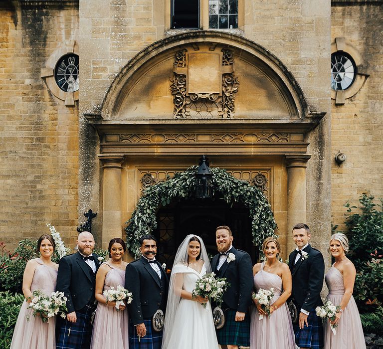 Wedding party portrait with bridesmaids in pale pink dresses, and groomsmen in tartan kilts at Foxhill Manor with foliage arch decor 