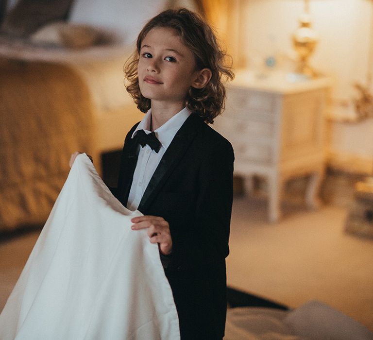 Page boy wears black tie and holds train of Suzanne Neville wedding gown