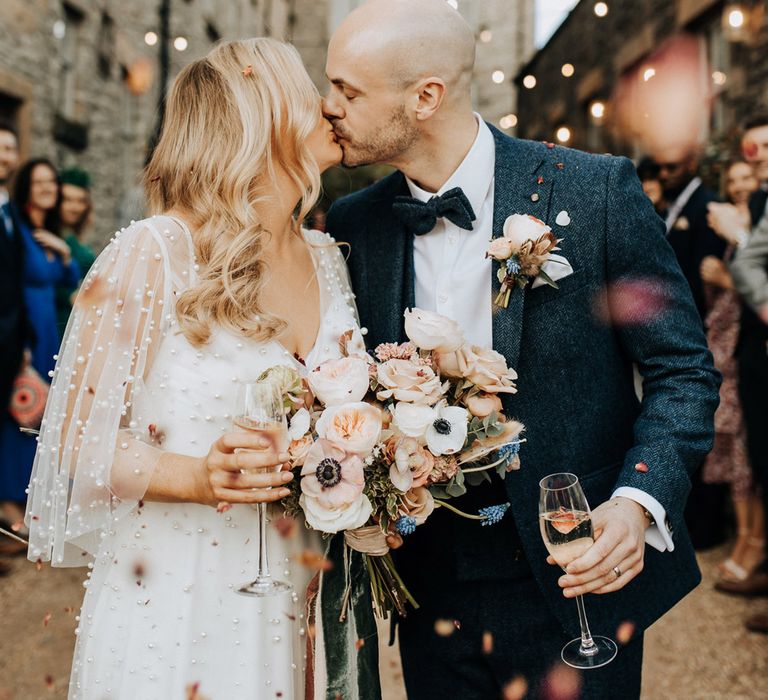 Bride with blonde hair and wedding dress with short sleeves encrusted with pearls holding pastel wedding bouquet kisses groom in navy tweed wedding suit with buttonhole