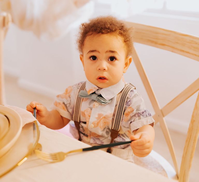 Page boy in a patterned shirt with bow tie and braces sitting at the top table with gold cutlery 