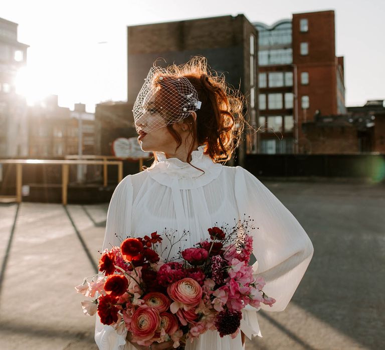 Bride in a high neck wedding dress with long sleeves wearing a birdcage veil with pearls on holding a romantic red bouquet during golden hour 