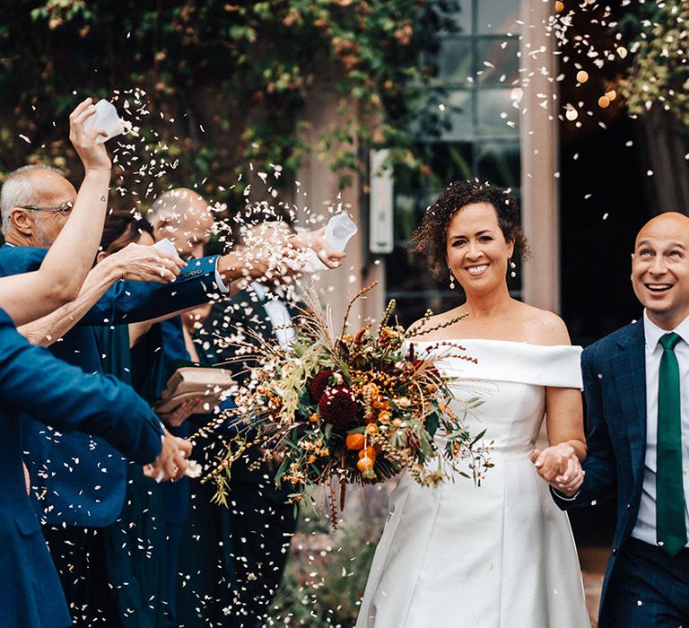 Bride in off the shoulder wedding dress with red and orange autumnal wedding bouquet has confetti exit with groom in blue suit