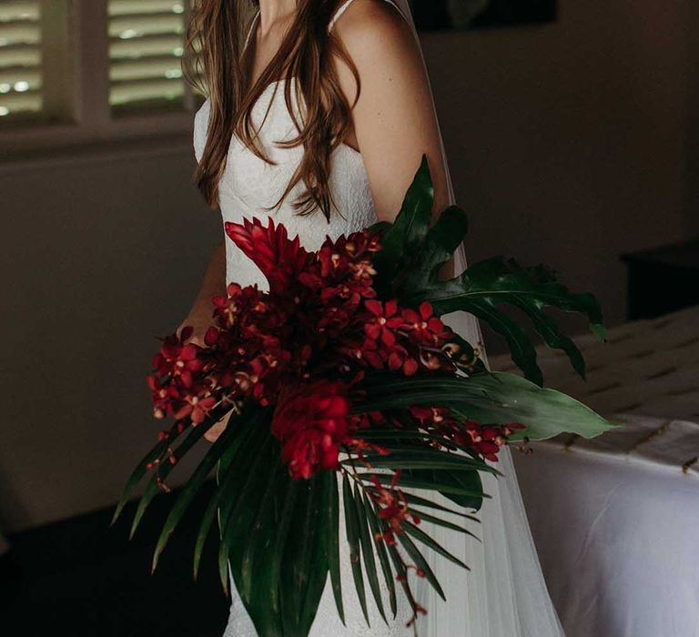 Bride in lace wedding dress with half up half down hair and veil holds bright red tropical flower wedding bouquet 