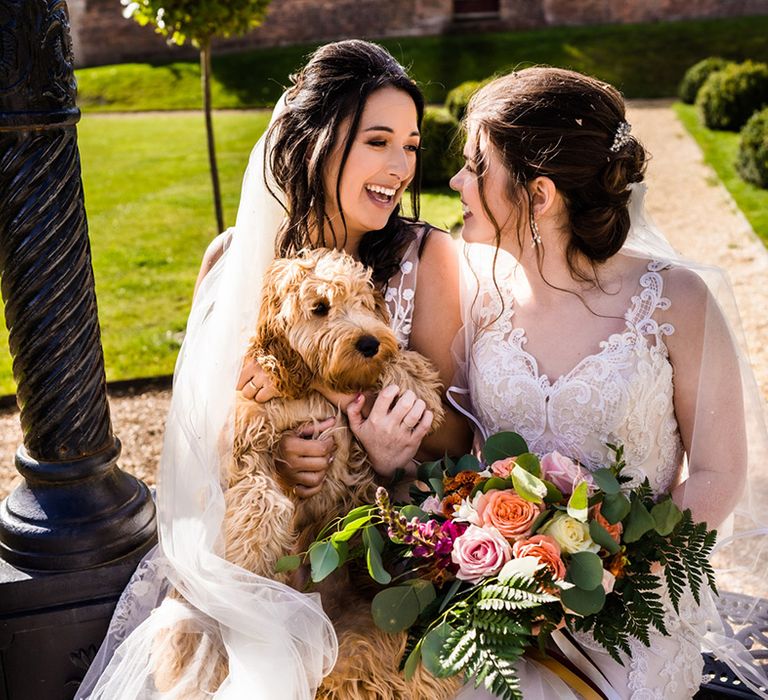 Brides in lace wedding dresses sit together with their pet dog and colourful wedding bouquet 