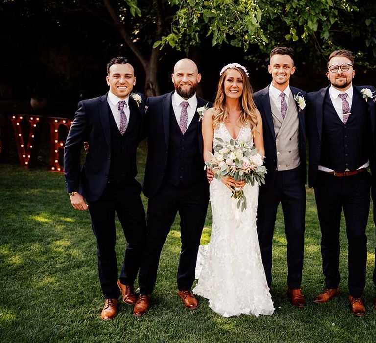 Bride in lace wedding dress and headband stands with groom and groomsmen in matching suits and ties with brown shoes