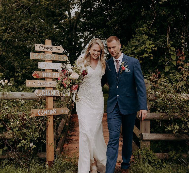 Bride in beaded wedding dress and groom in blue suit walk next to their DIY wedding sign decor