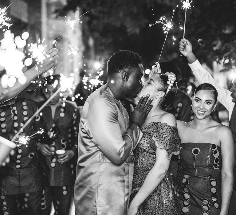 Bride and groom share a passionate kiss as their guests wave sparklers around 