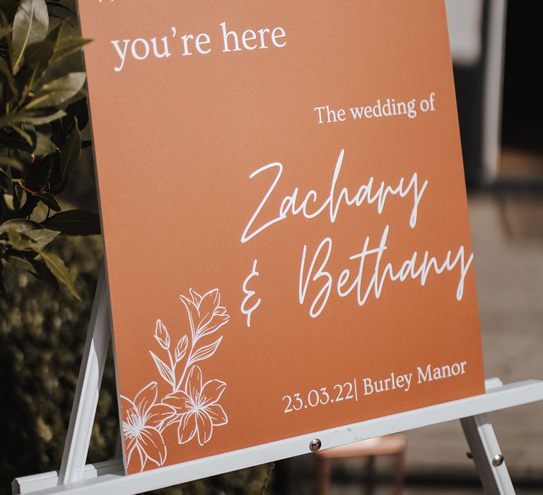 Orange rust coloured welcome wedding sign with flower design and white font