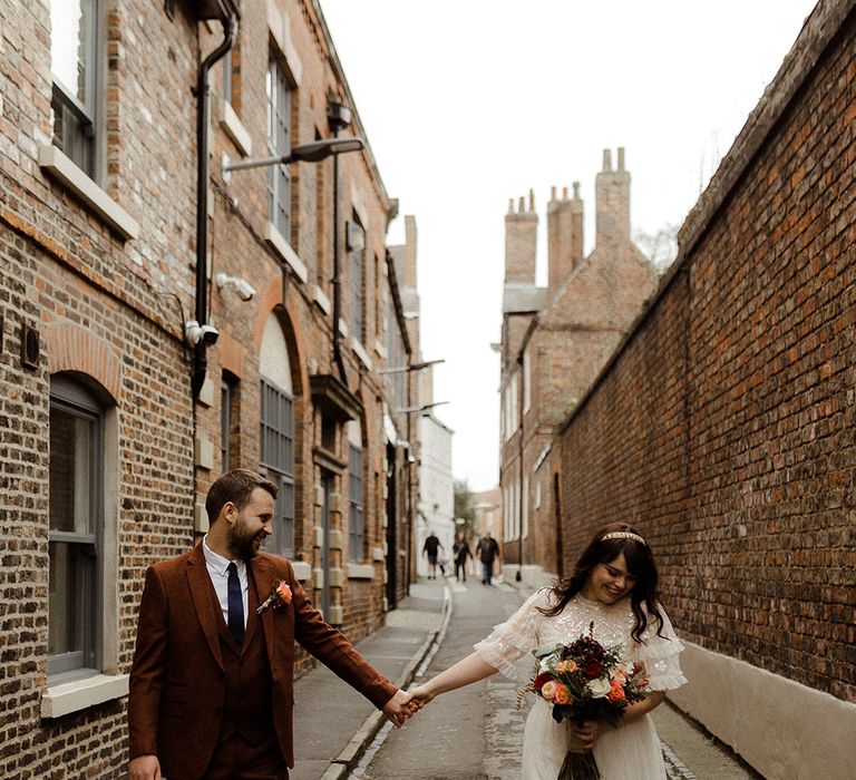 Bride and groom smile holding hands as they walk down an alley together in York